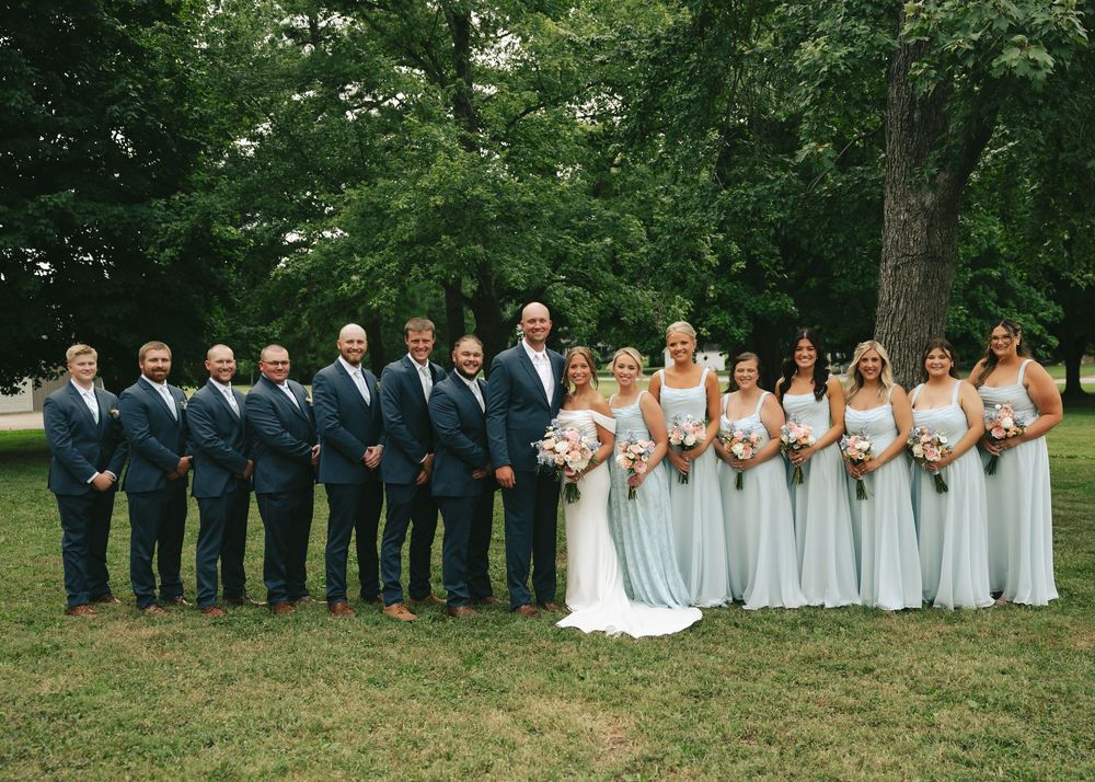 Wedding party posing outdoors with groomsmen in navy suits and bridesmaids in mint gowns flanking bride and groom.