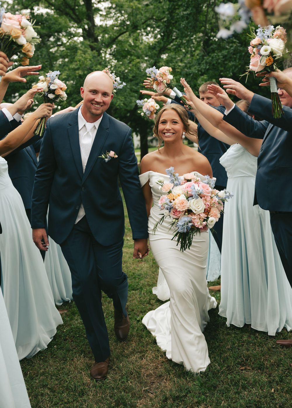 Smiling bride and groom walk through arch of bouquets held by wedding party in blue attire outdoors.