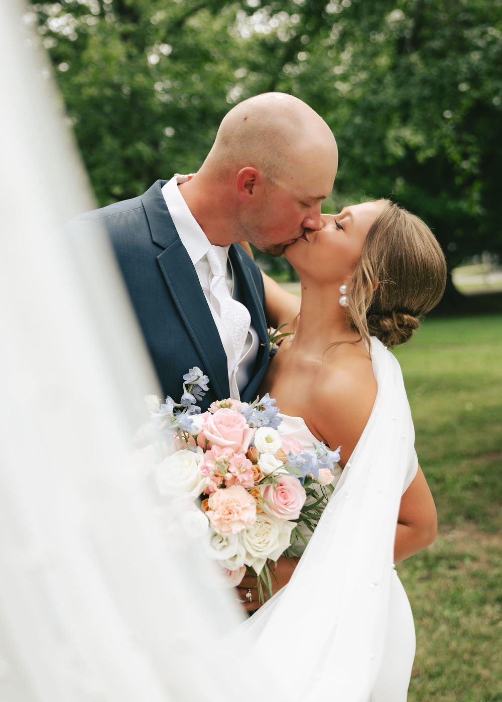 Bride and groom share a romantic kiss outdoors, bride holding a pastel bouquet of pink, white, and blue flowers.