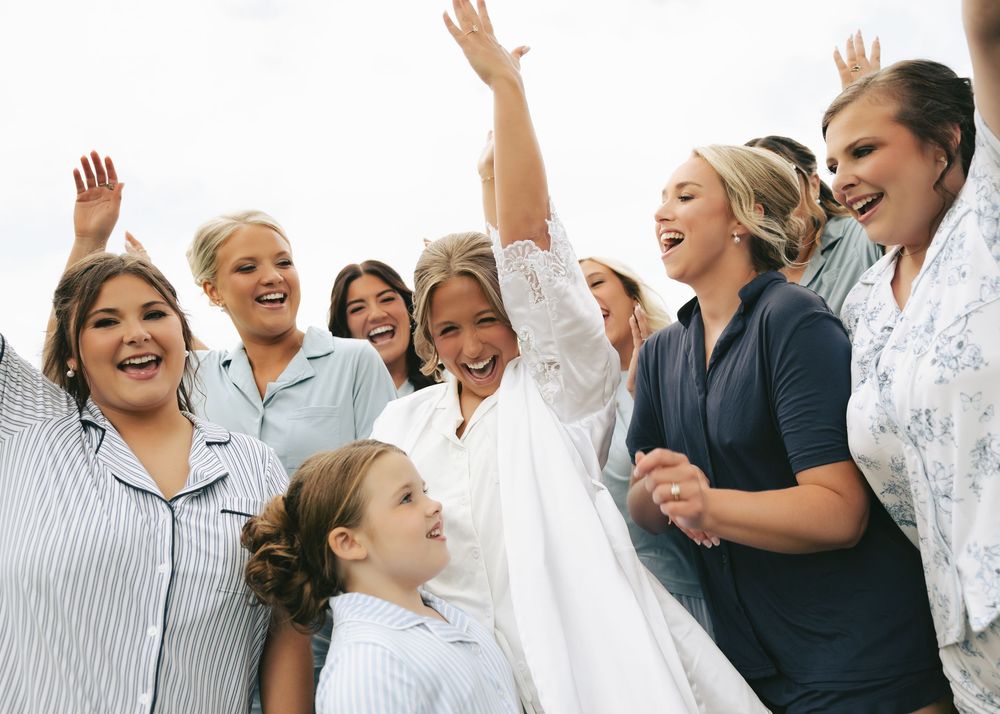 Joyful bride in white dress celebrating with bridesmaids and flower girl, arms raised in excitement.