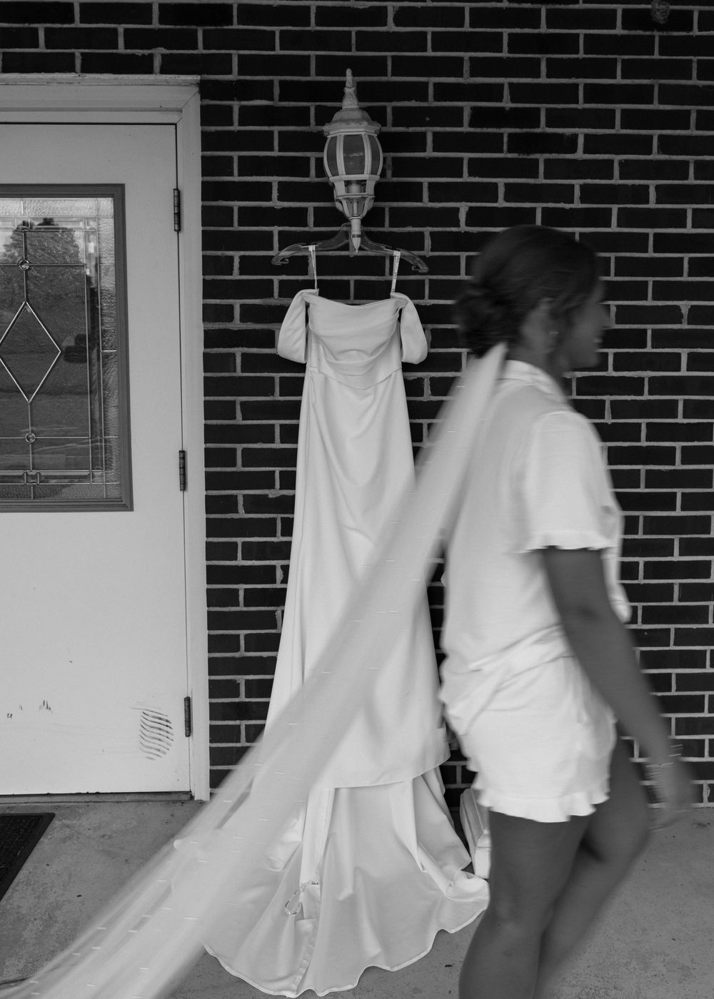 Black and white photo of a woman handling a white wedding dress hung on a brick wall outside a door.