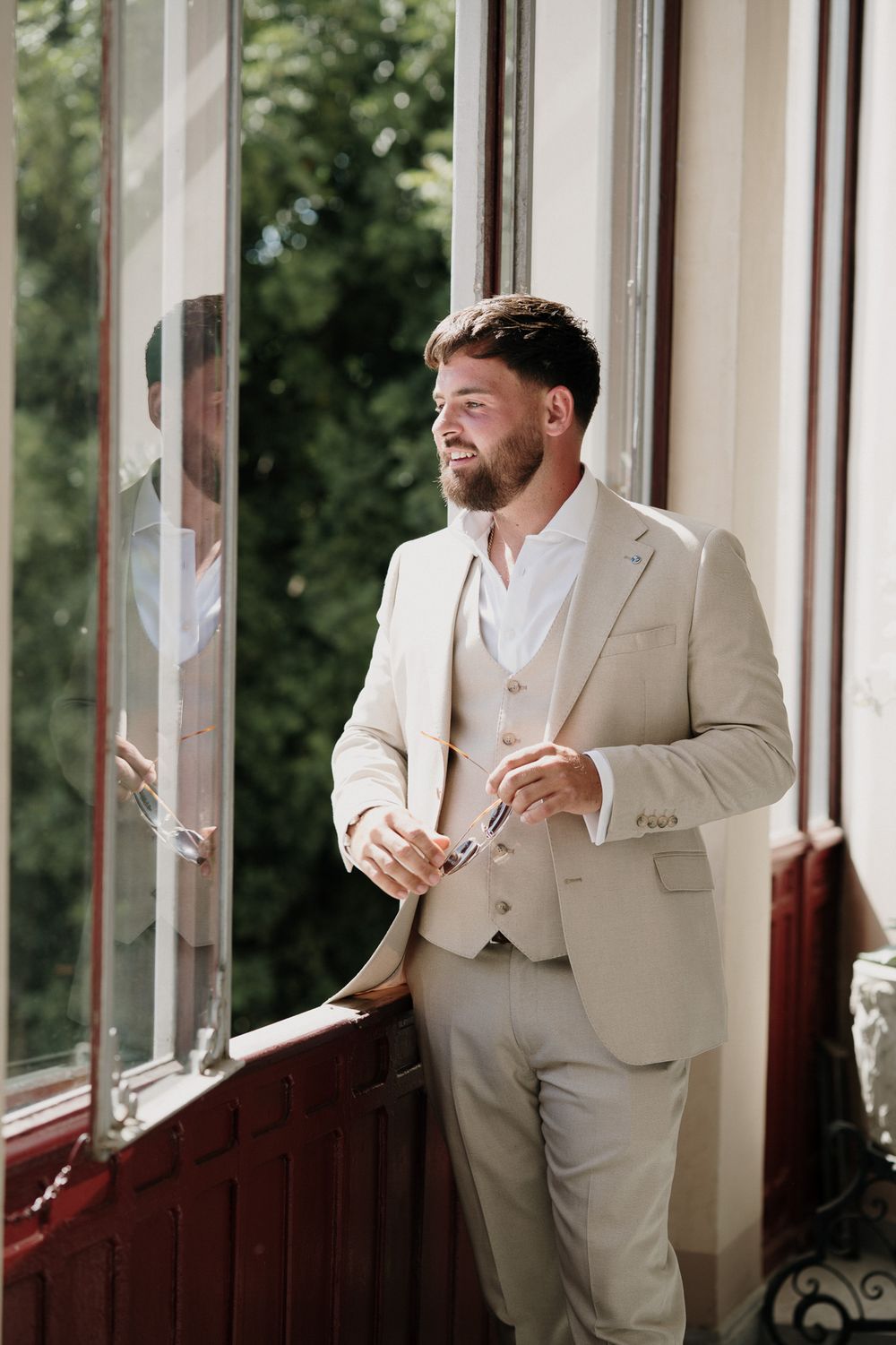 An elegant groom in a beige suit looks out the window at Villa Nobel in Sanremo.