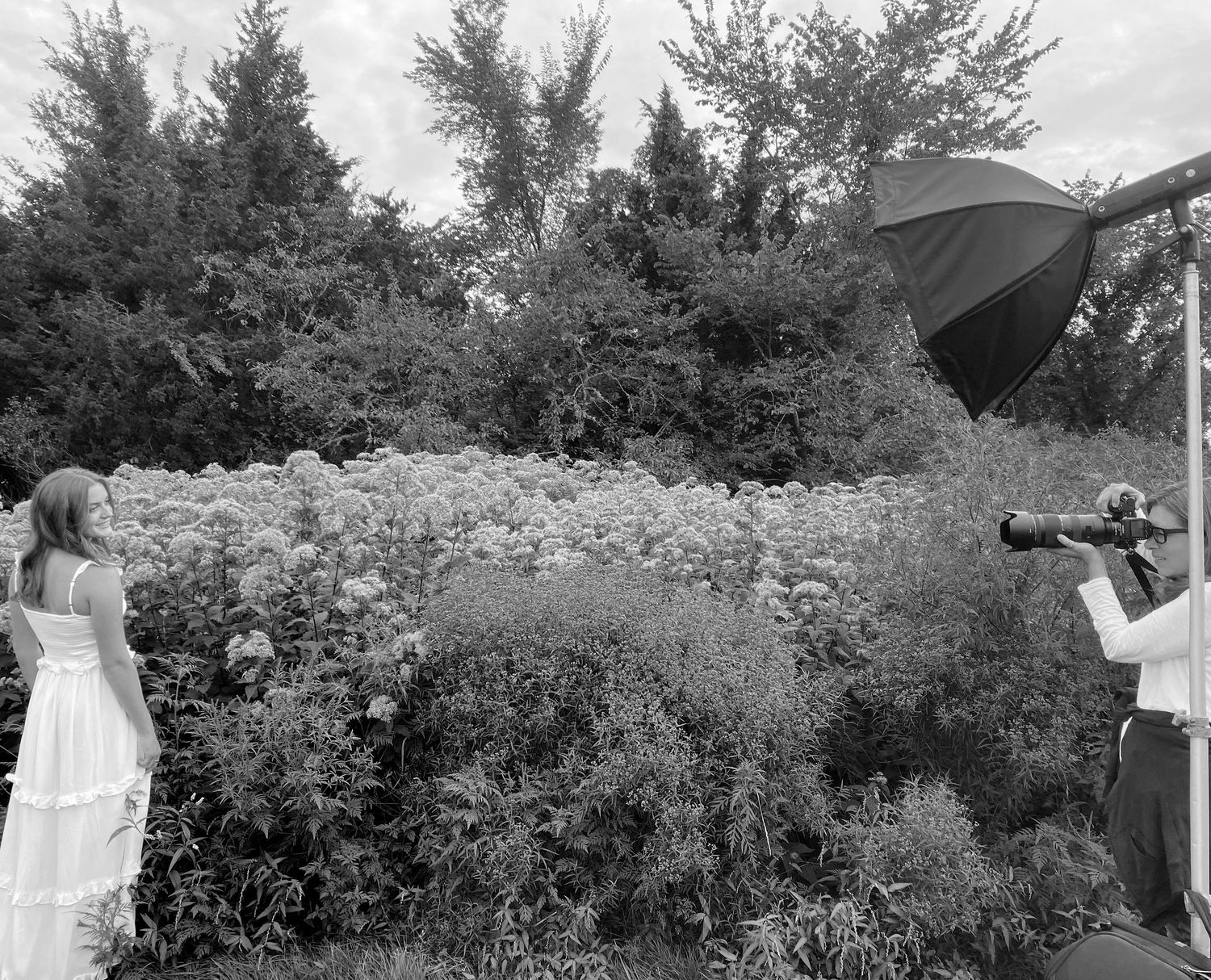 A photographer with lighting equipment captures a photo of someone in a white dress against a garden backdrop.