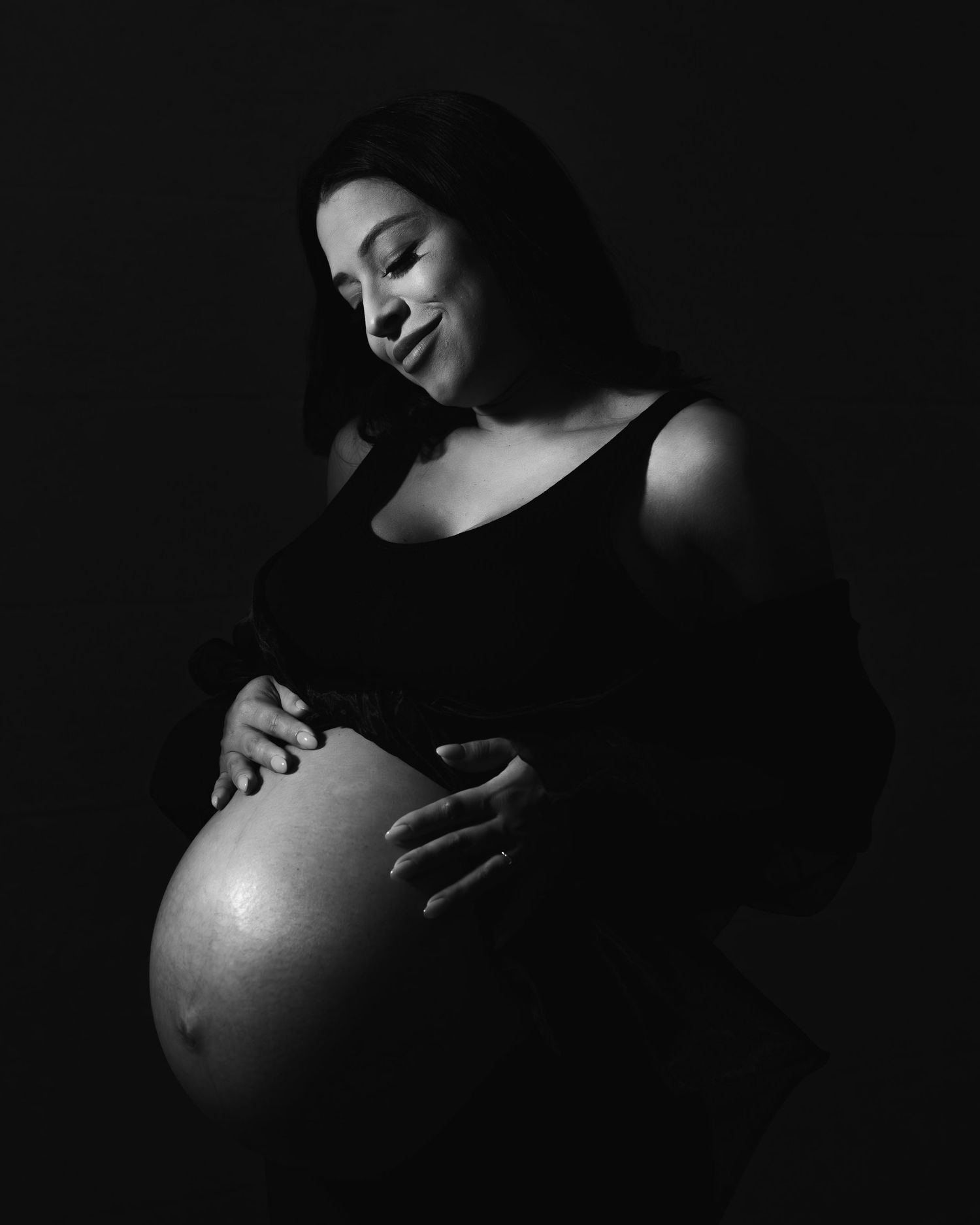 Black and white maternity photo of a pregnant woman cradling her bare belly, smiling softly against a dark background.