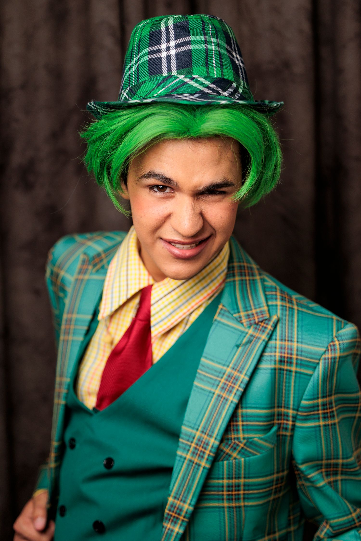 A young male performer in a green plaid suit and matching hat with bright green wig grins mischievously.