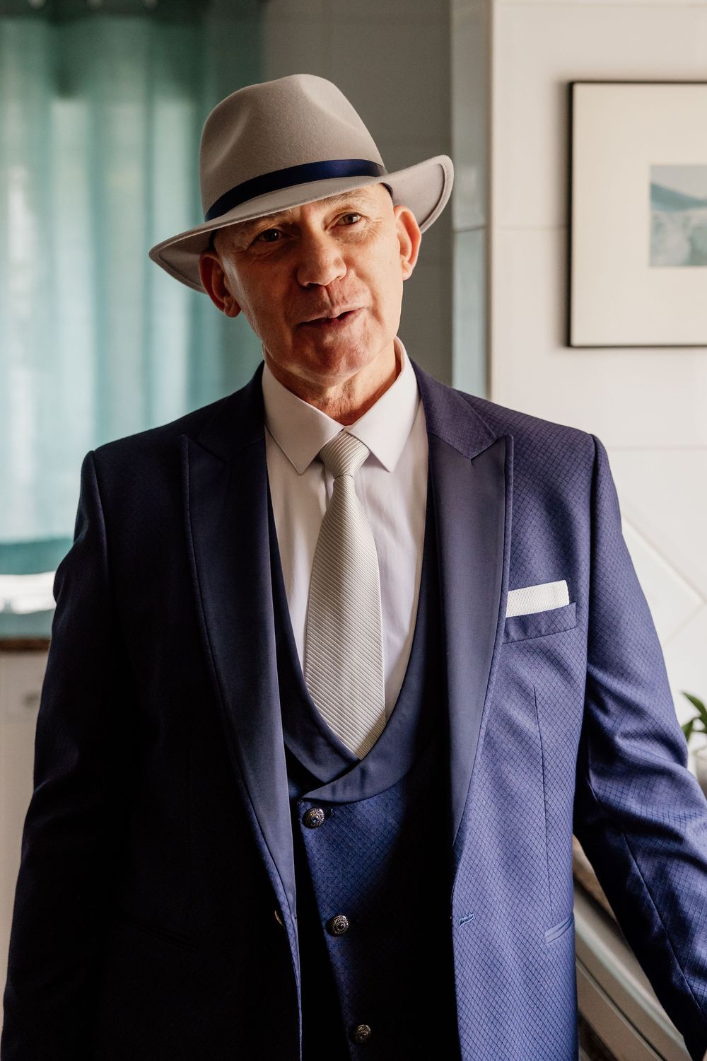 A well-dressed older man wearing a suit, hat, and tie stands in a room with a window in the background.