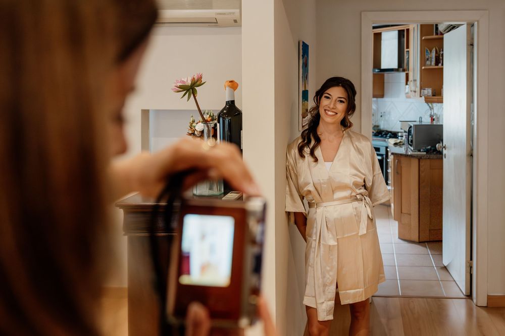 A woman in a beige robe stands in a kitchen, holding a phone and smiling.