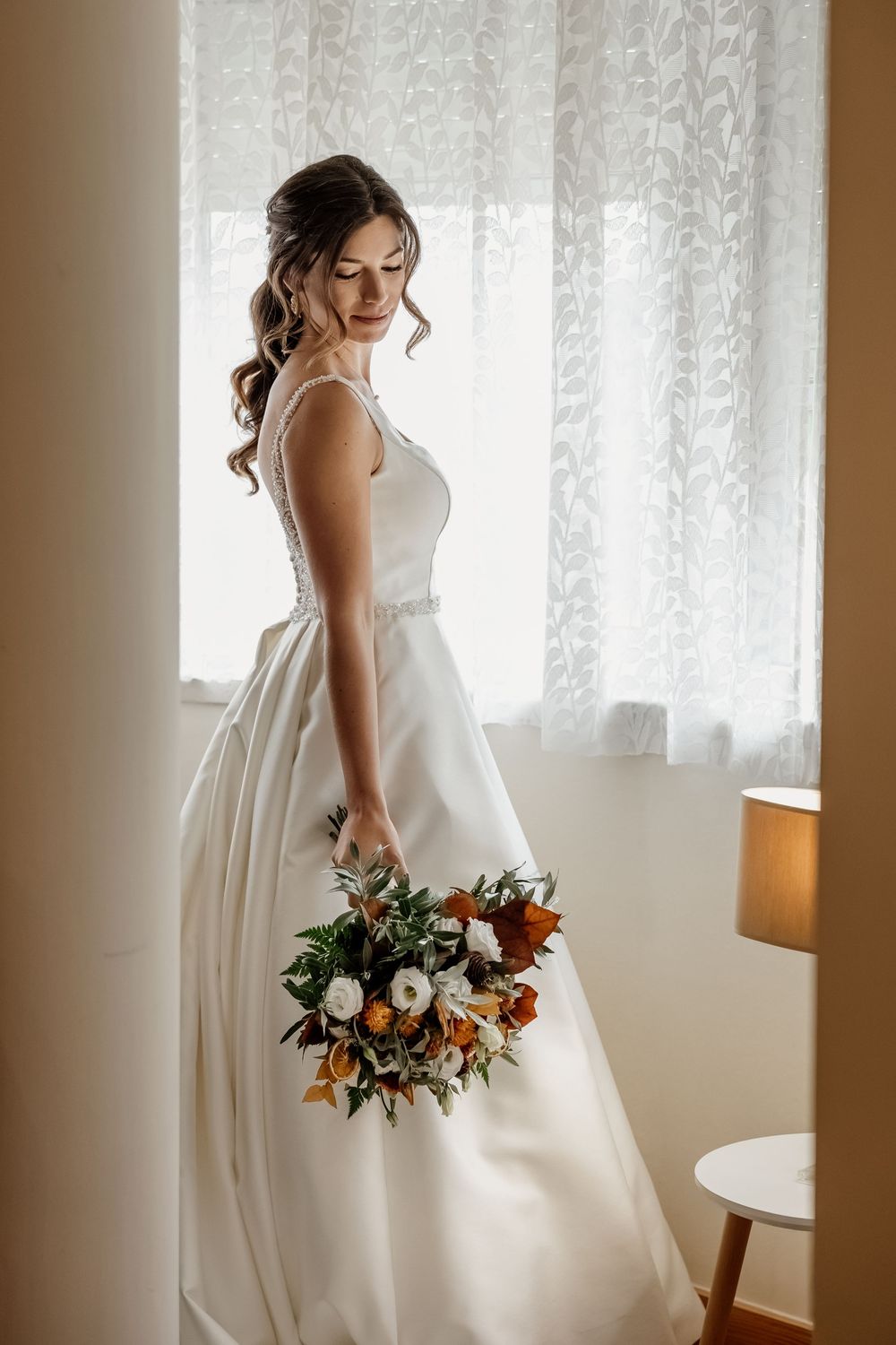 A bride stands in a room, holding a bouquet of flowers, wearing a white wedding dress and looking away from the camera.