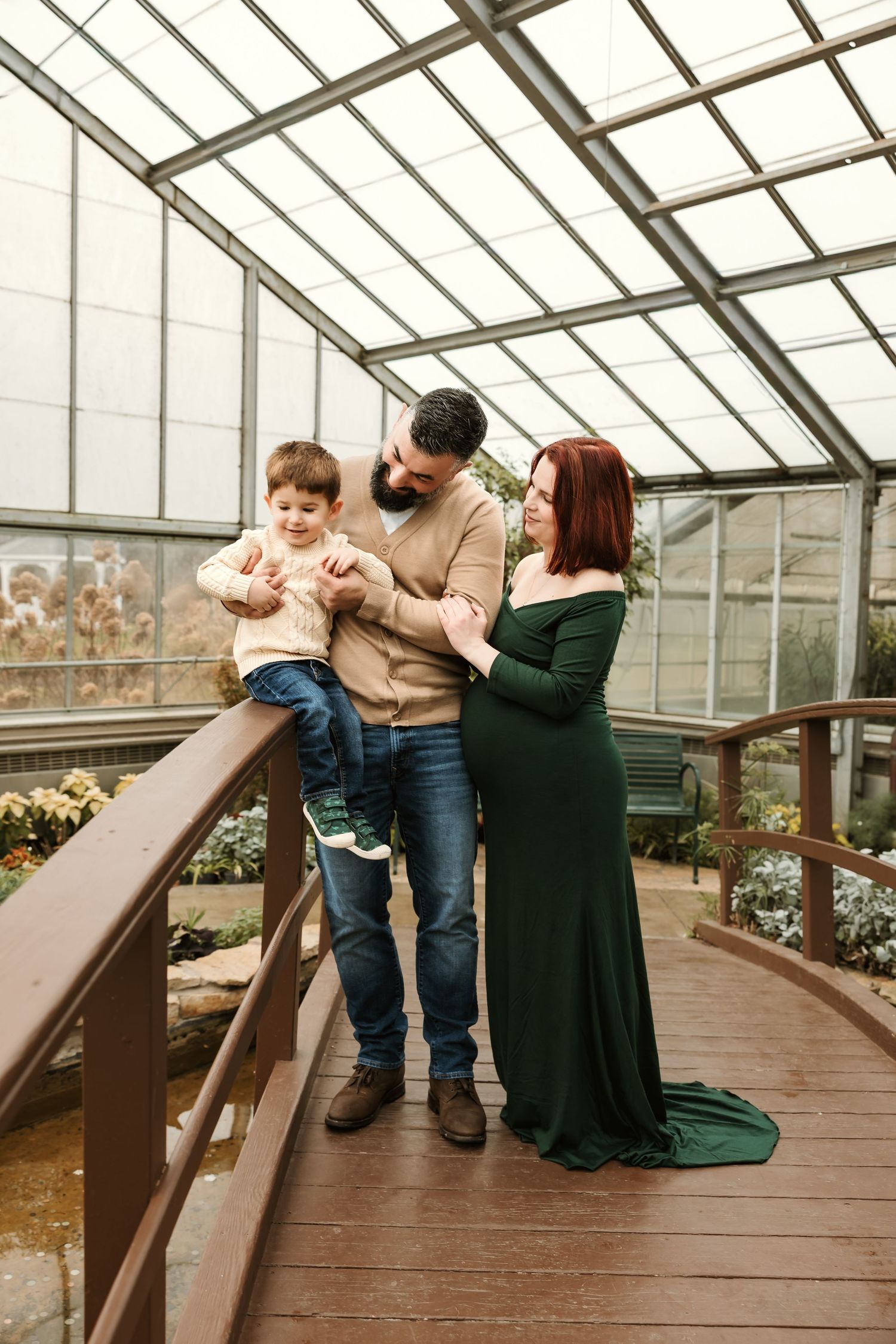 Family with toddler son posing on wooden bridge inside glass greenhouse conservatory.