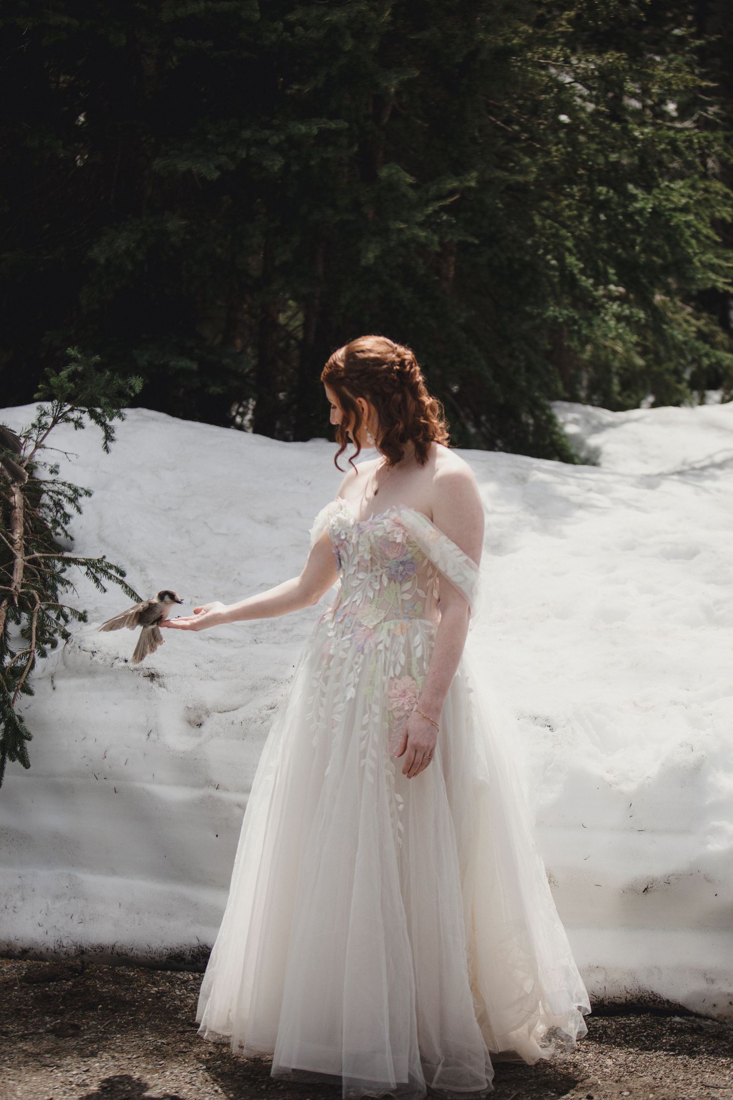 Two people in matching white floral wedding dresses stand together in the snow during a winter photo session.