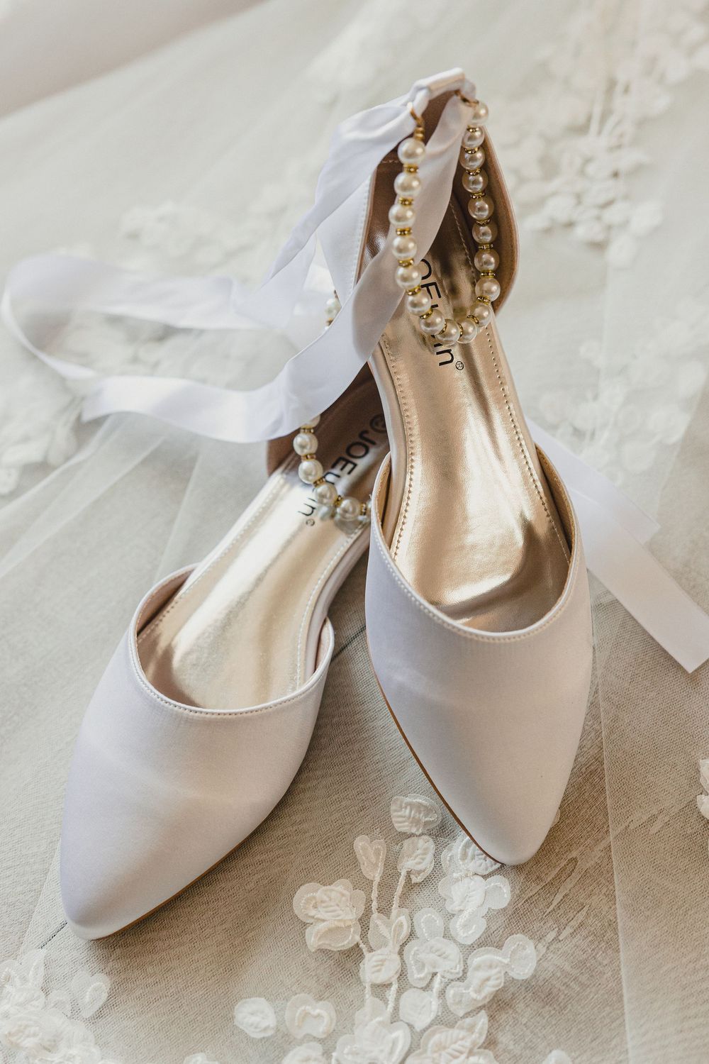 Elegant white satin bridal shoes with pearl embellishments displayed on a textured white surface.