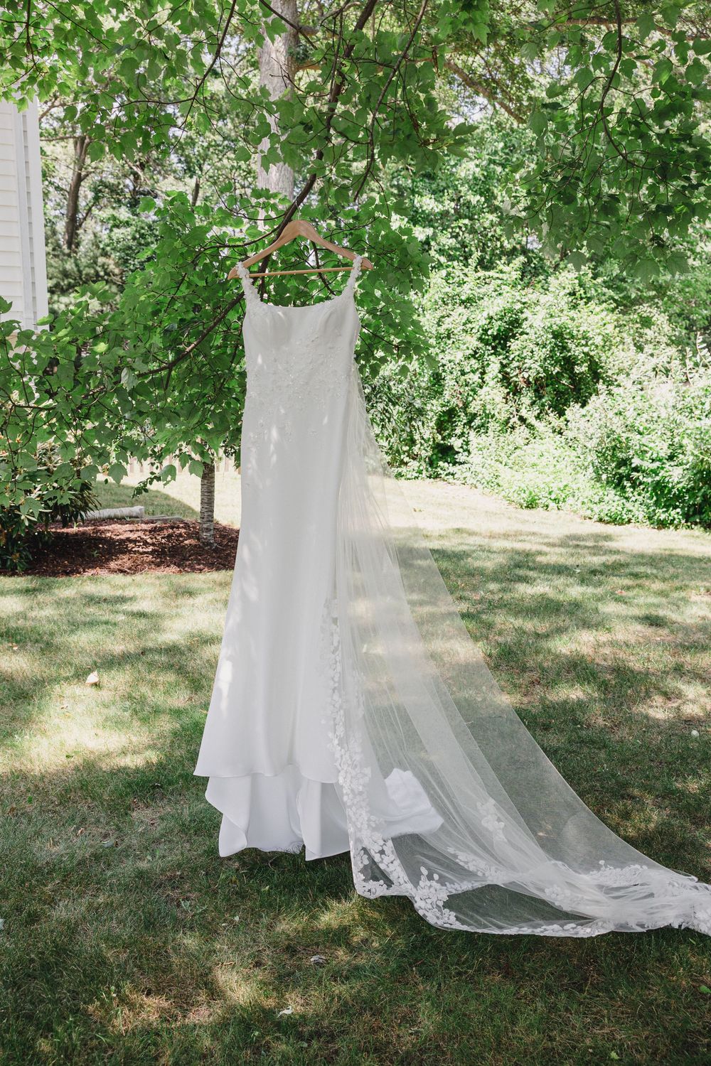 White wedding dress hanging outdoors against a natural green garden backdrop with sunlight filtering through trees.