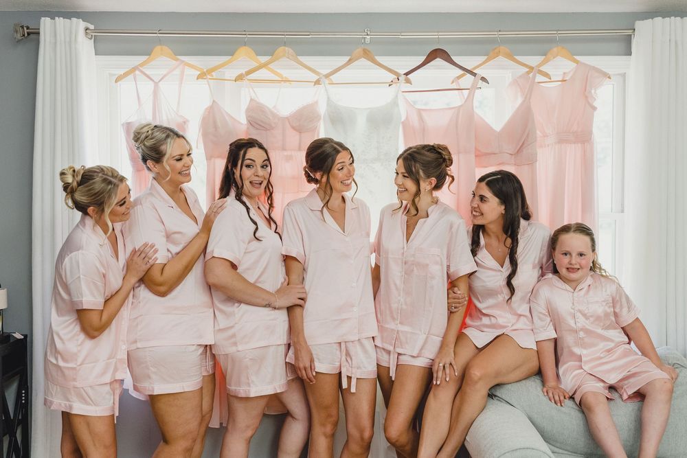 Bridesmaids in matching pink robes gather by a window during wedding preparations.