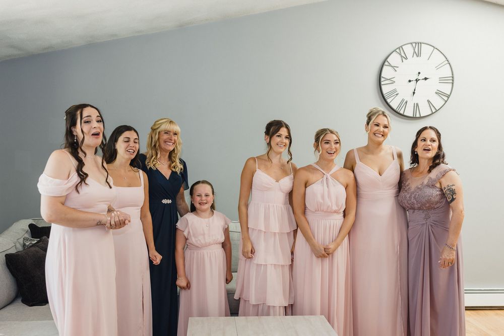Bridesmaids in pink and purple dresses gather together in a bright room with a wall clock.