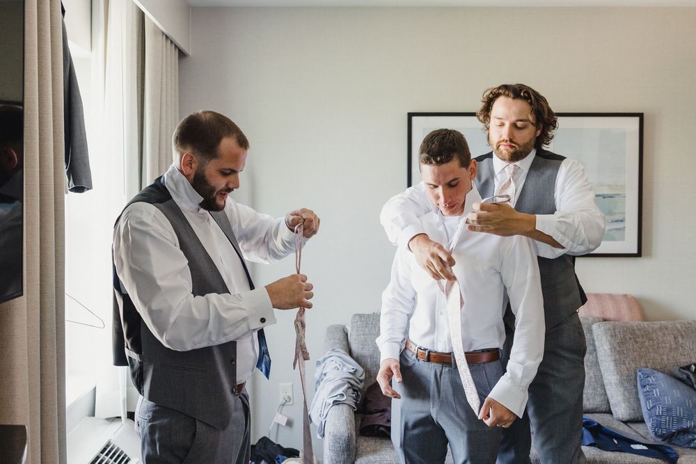 Three people getting dressed in formal attire while standing in a bright room with white walls and framed artwork.