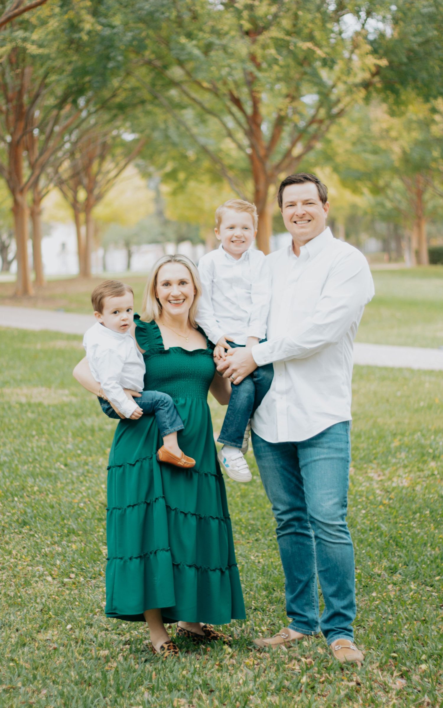 Family portrait outdoors in a park with tree-lined path, wearing coordinated white and emerald green outfits.