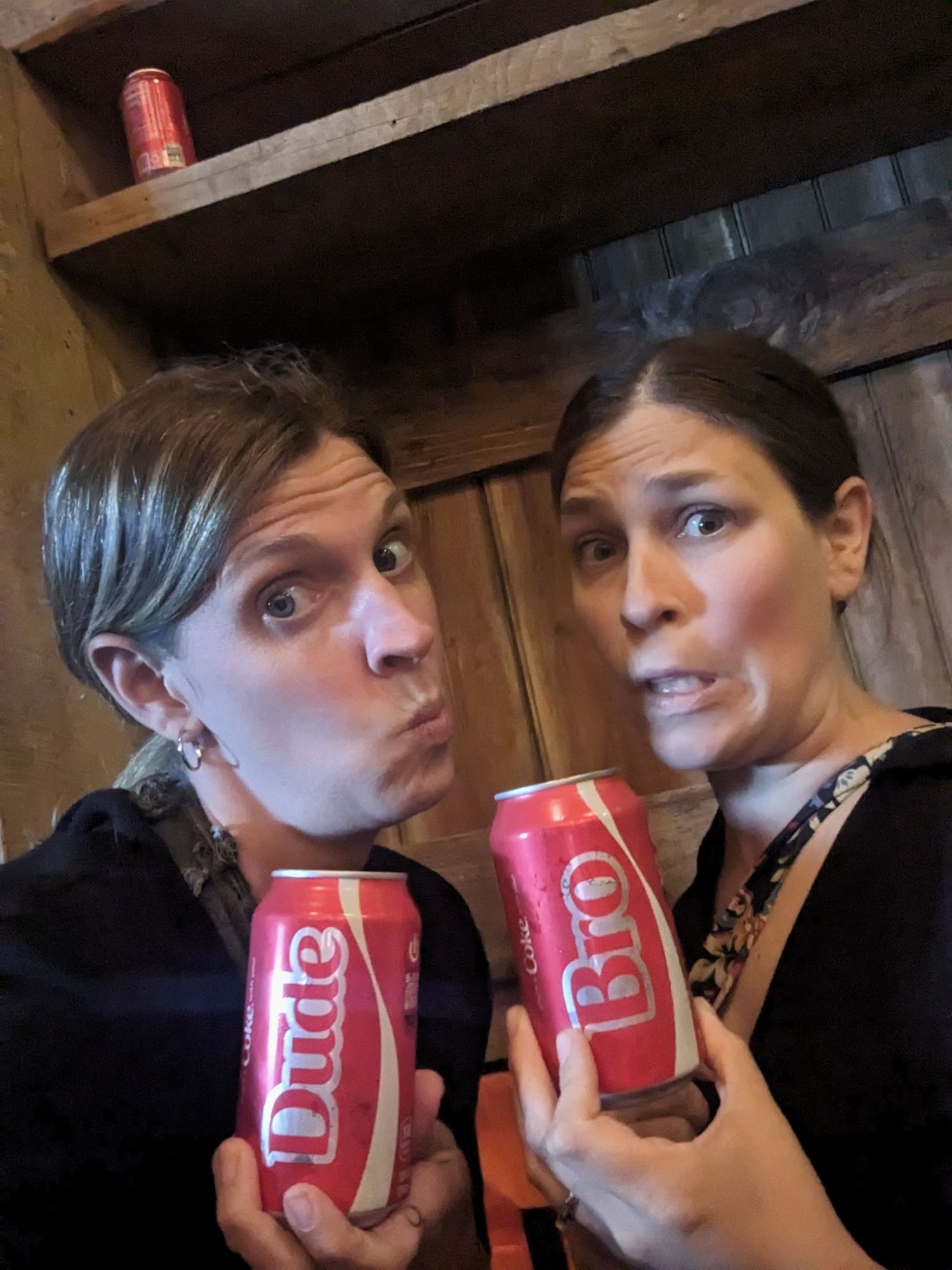 Two people make silly faces while holding personalized Coca-Cola bottles with names on them in a dimly lit room.