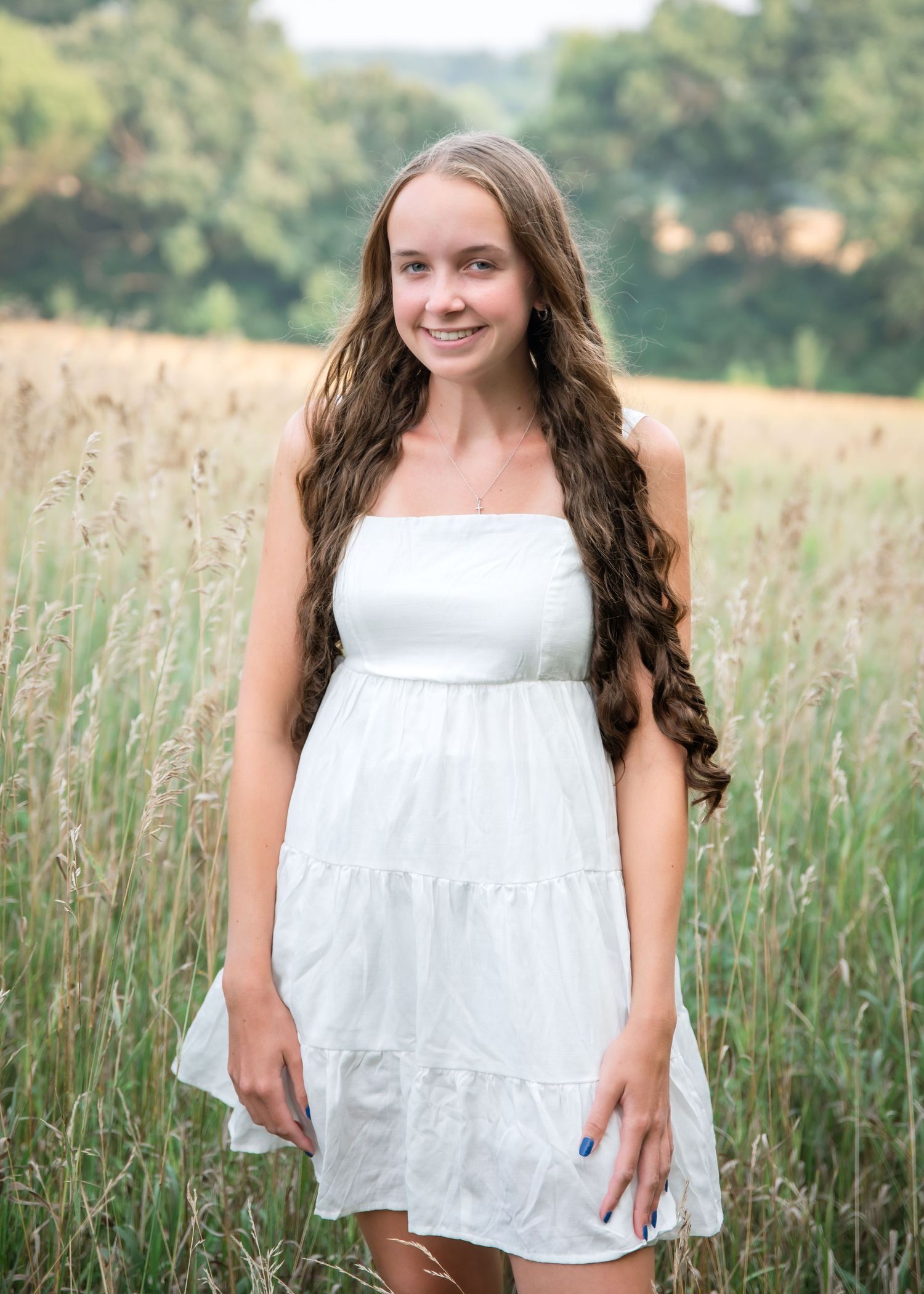 A person in a white dress sits joyfully in a tall grass meadow on a summer day with trees in the background.