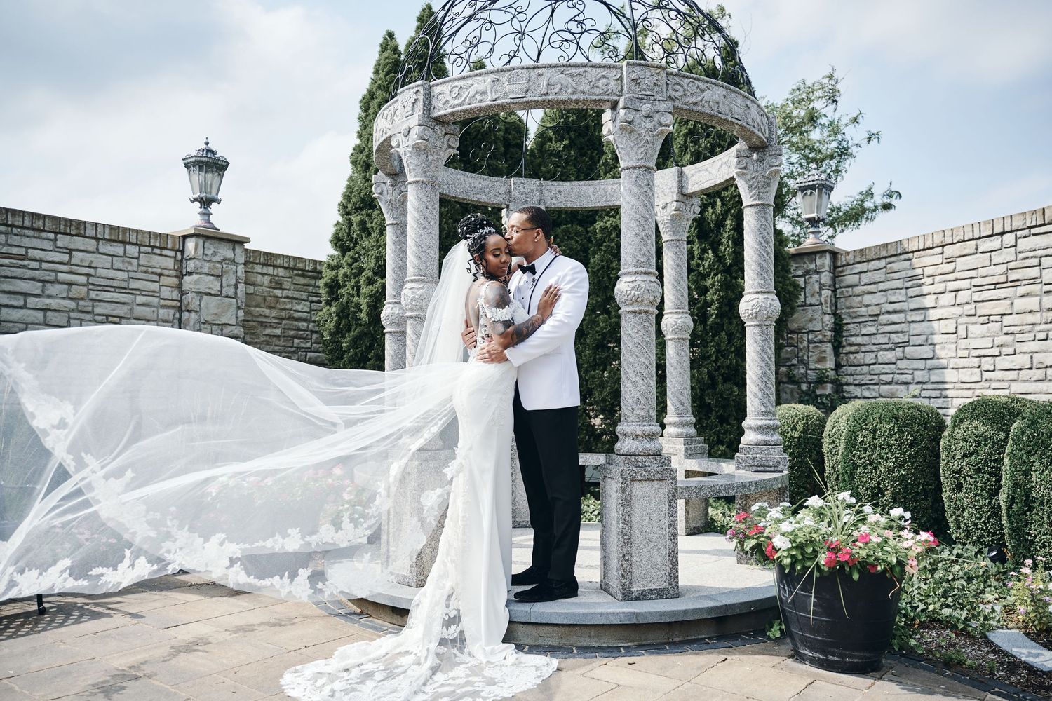 A romantic wedding ceremony takes place under an elegant stone gazebo surrounded by lush greenery and flowers.