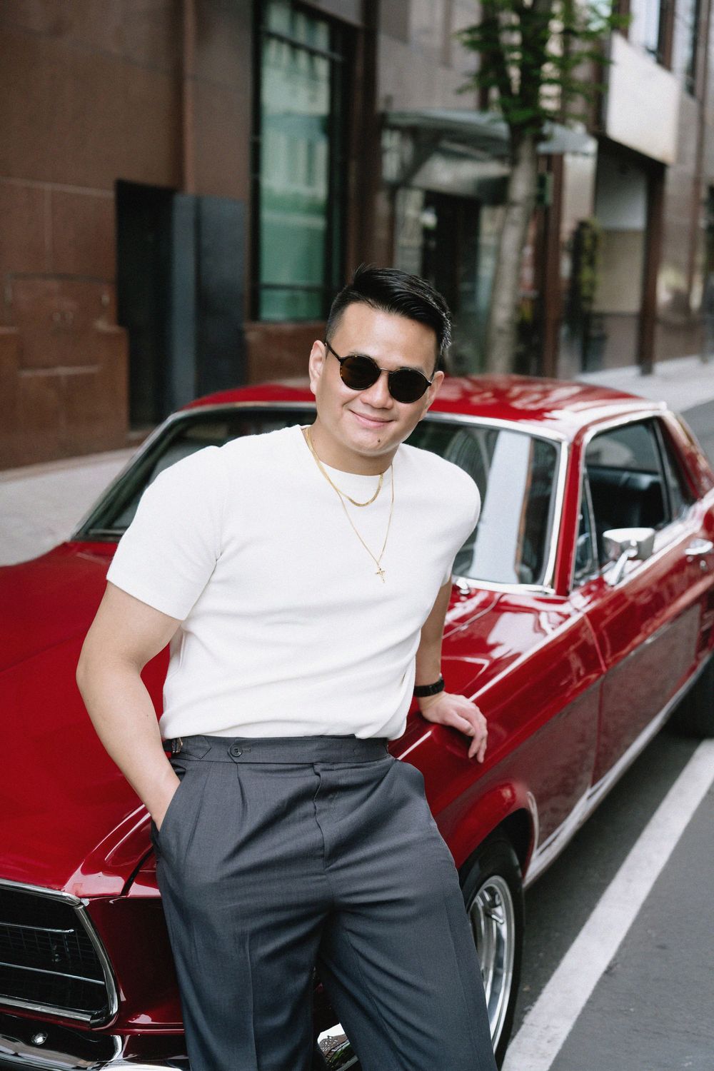 Confident man in white t-shirt and sunglasses leaning against a classic red vintage car on city street.
