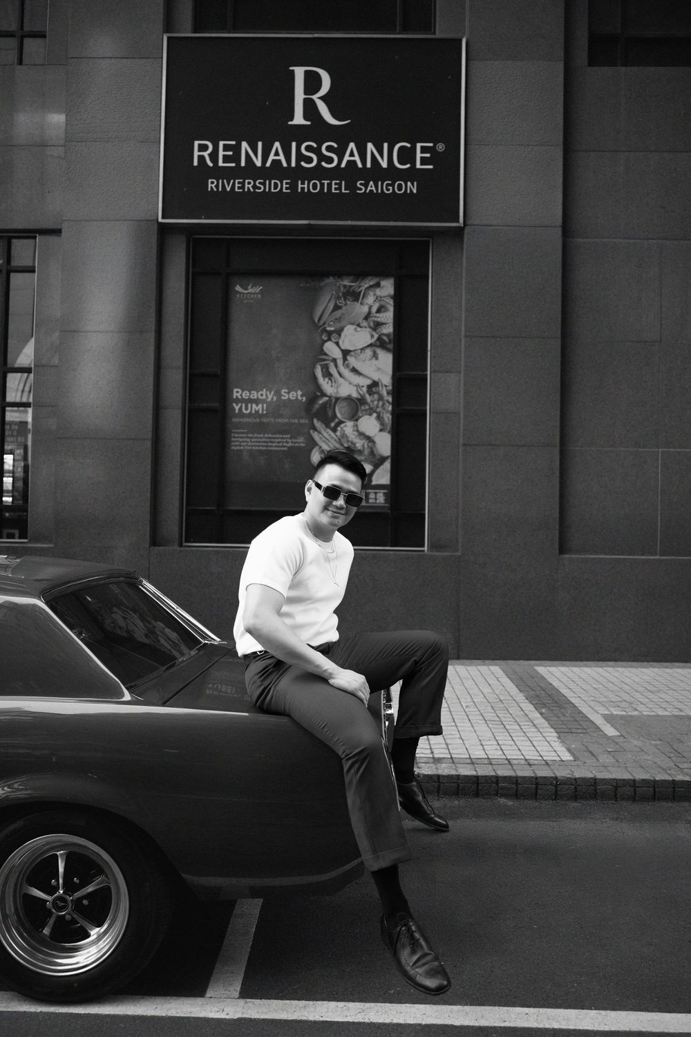 Stylish man in white t-shirt sitting on classic vintage car outside Renaissance Riverside Hotel Saigon.
