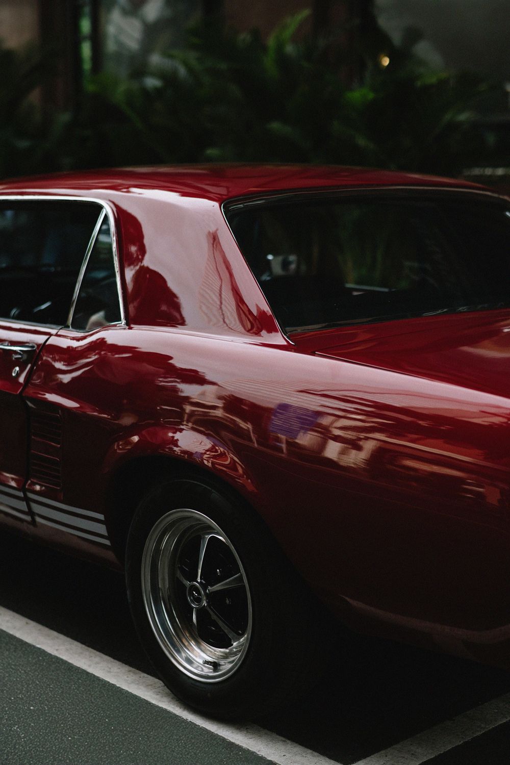 Sleek vintage red muscle car with chrome wheels parked against lush dark tropical garden background.