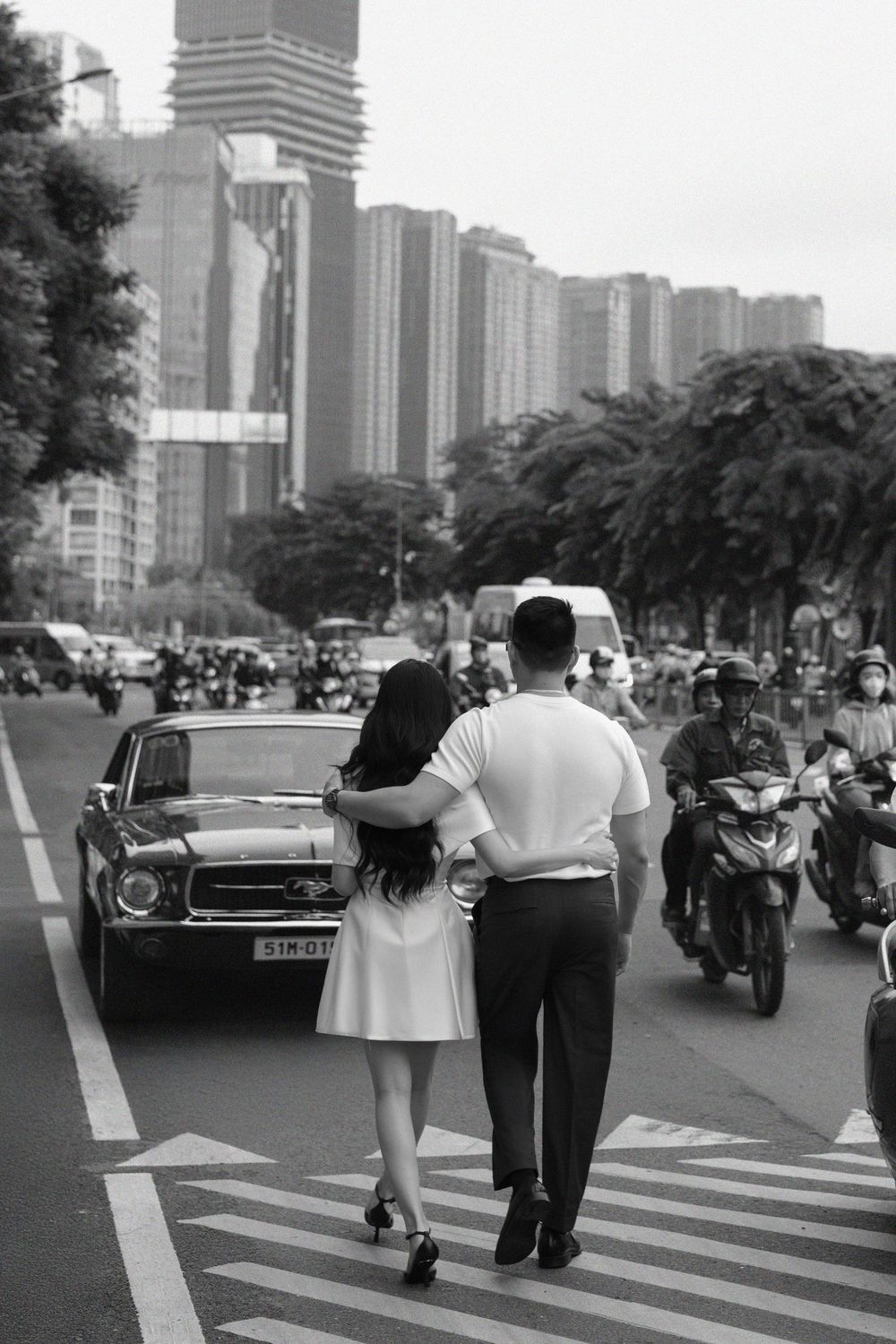 Black and white photo of a couple walking arm in arm across a crosswalk in a busy Asian city street.