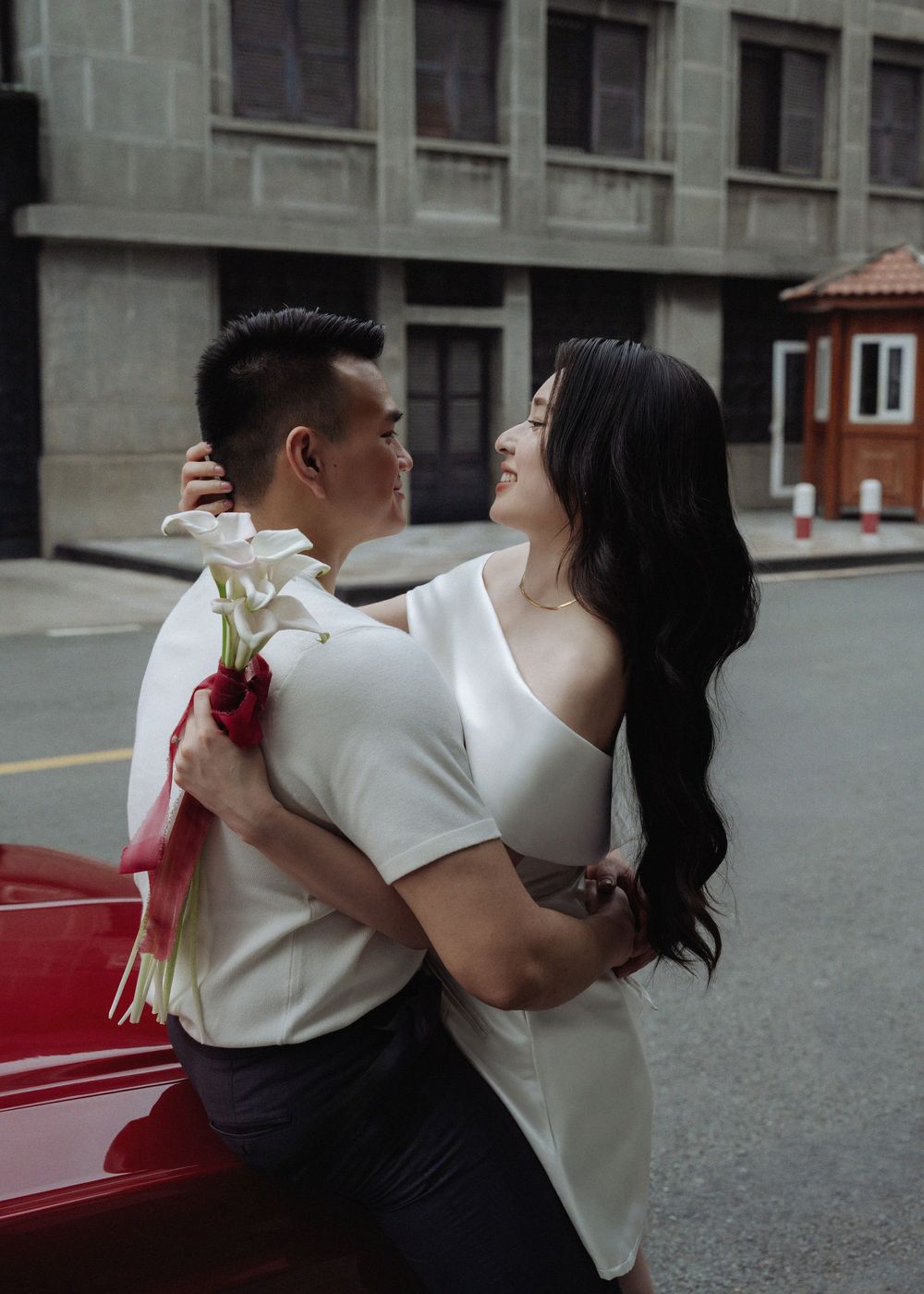 A couple embraces romantically against a red vintage car, woman holding white calla lilies.