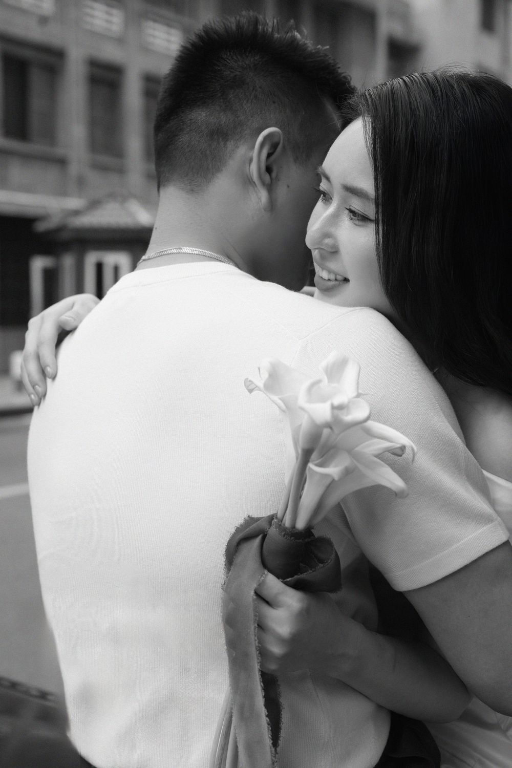 Black and white photo of a smiling woman embracing a man while holding a bouquet of calla lilies.