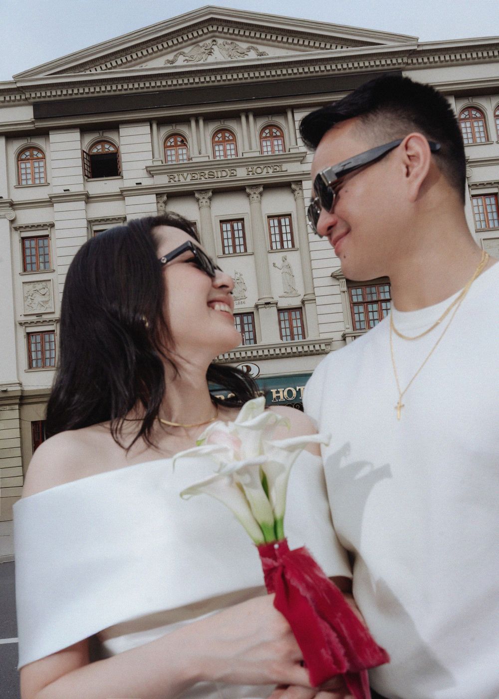 Couple in sunglasses gazing at each other holding white calla lilies in front of Riverside Hotel.