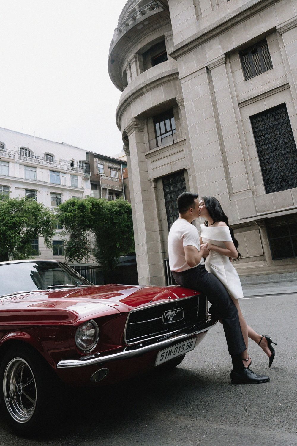 Couple sharing a romantic kiss while leaning against a red Ford Mustang in front of a stone building.