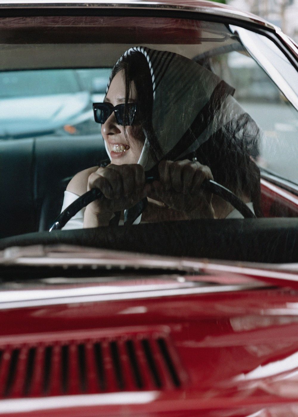 A stylish woman in sunglasses and headscarf drives a classic red car.