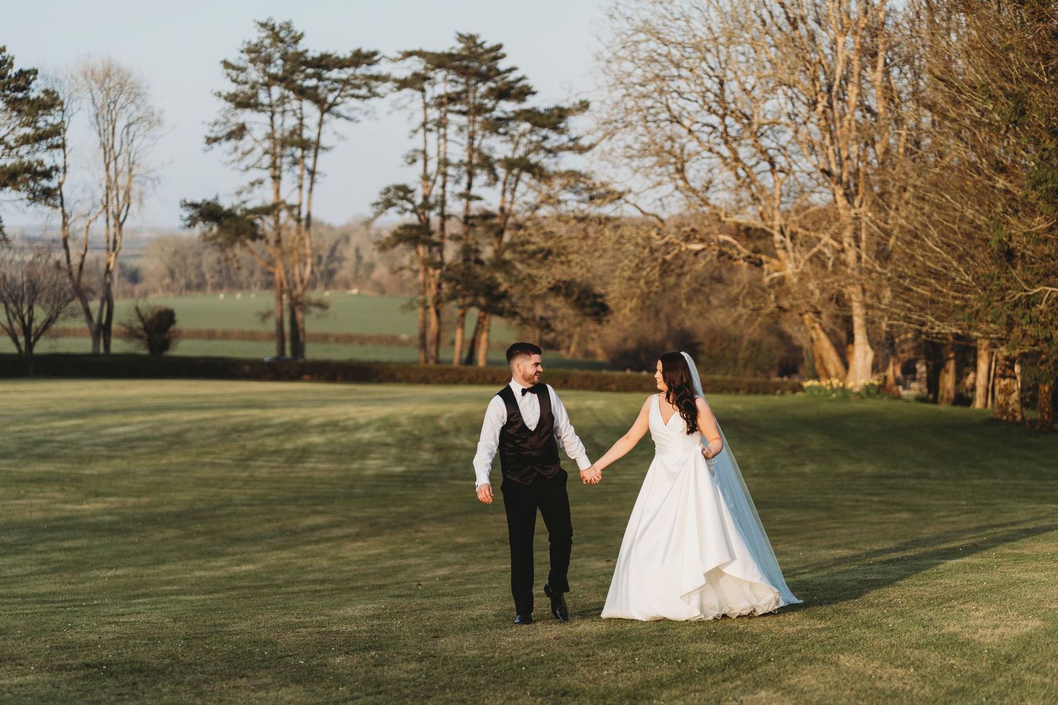 Bride and groom walking across countryside lawns during relaxed sunset wedding portraits in West Wales