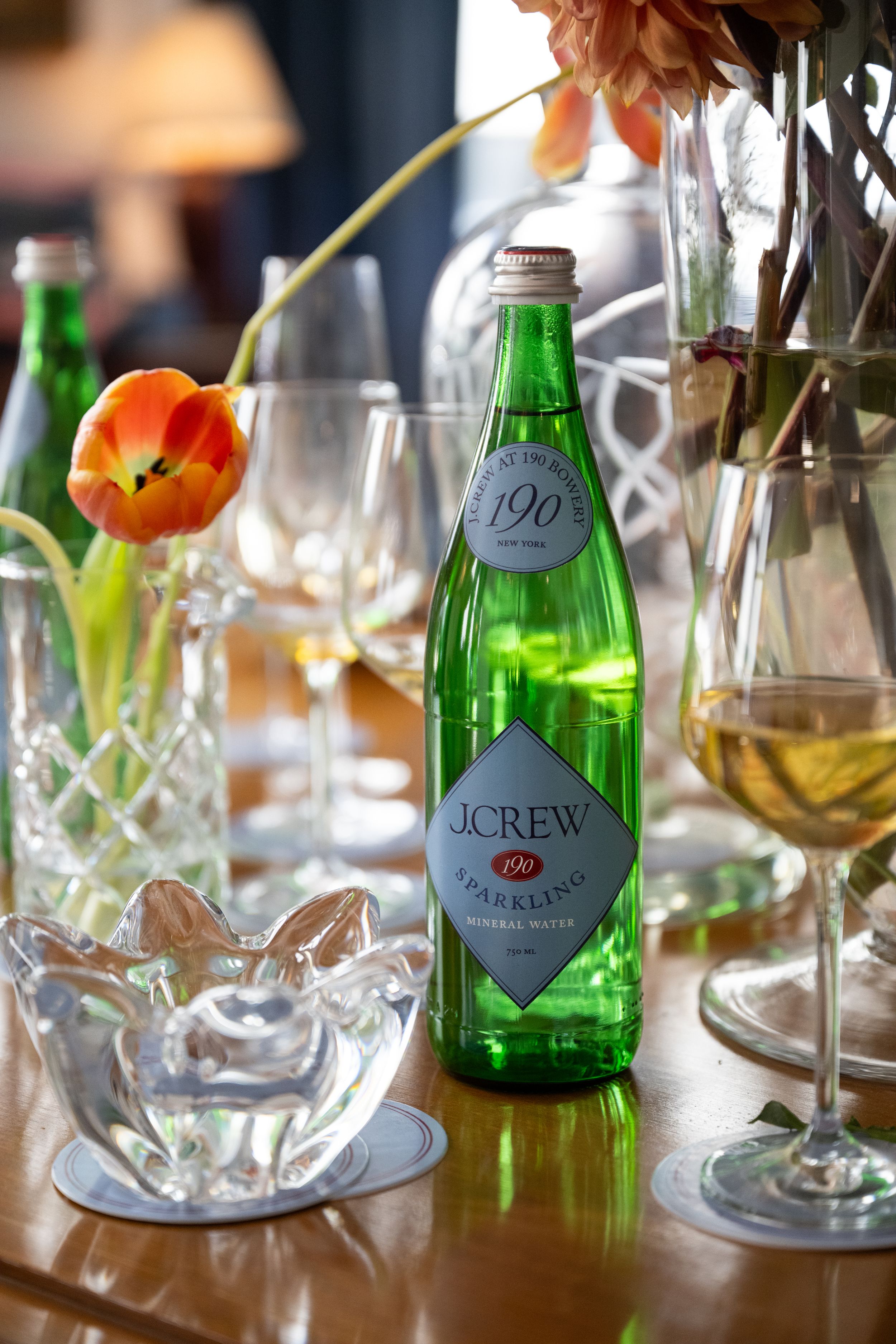 J.Crew sparkling mineral water green glass bottle on a wooden table with wine glasses and tulips.