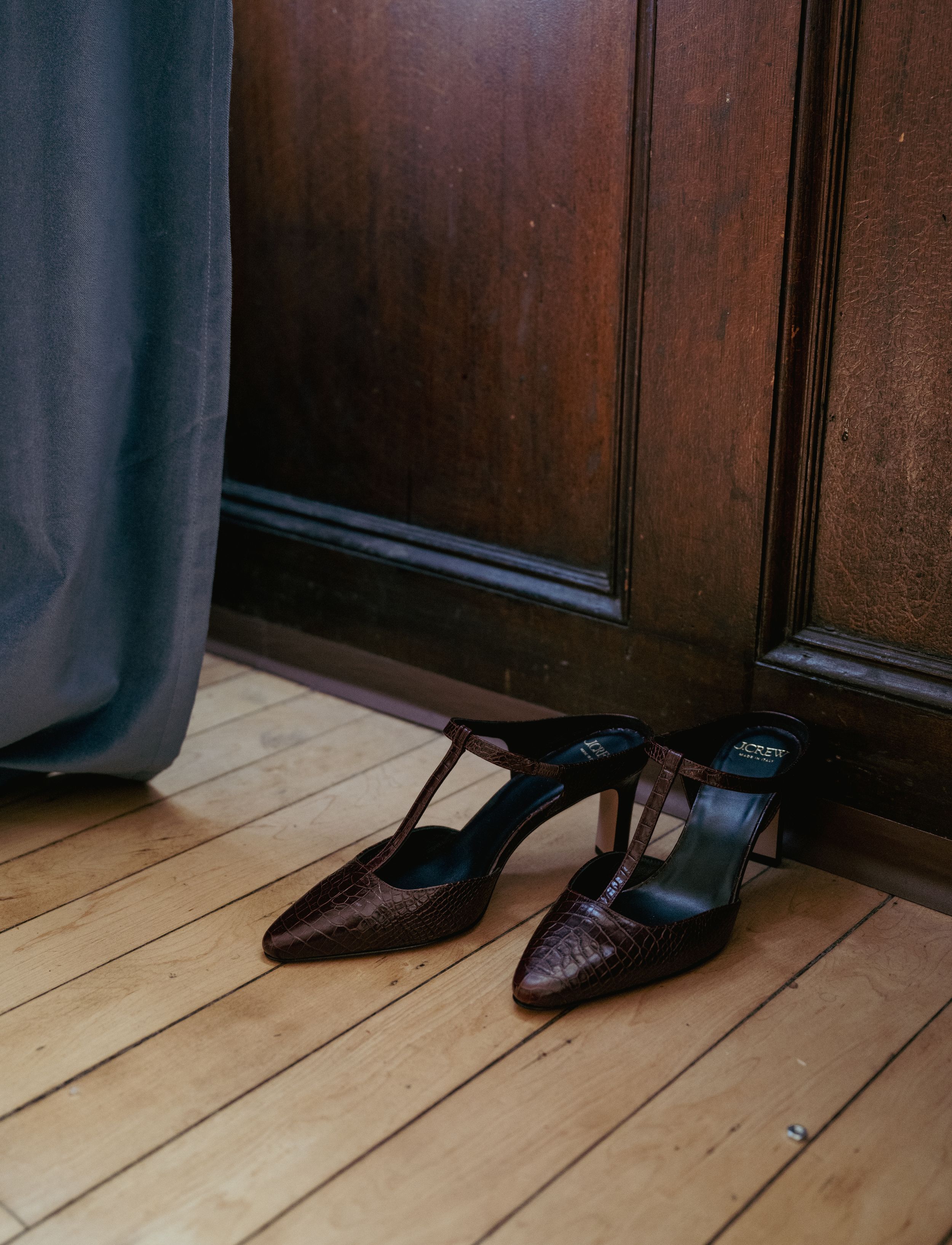 Brown croc-embossed stiletto mules with T-strap detail resting on hardwood floor beside wooden door and gray curtain.