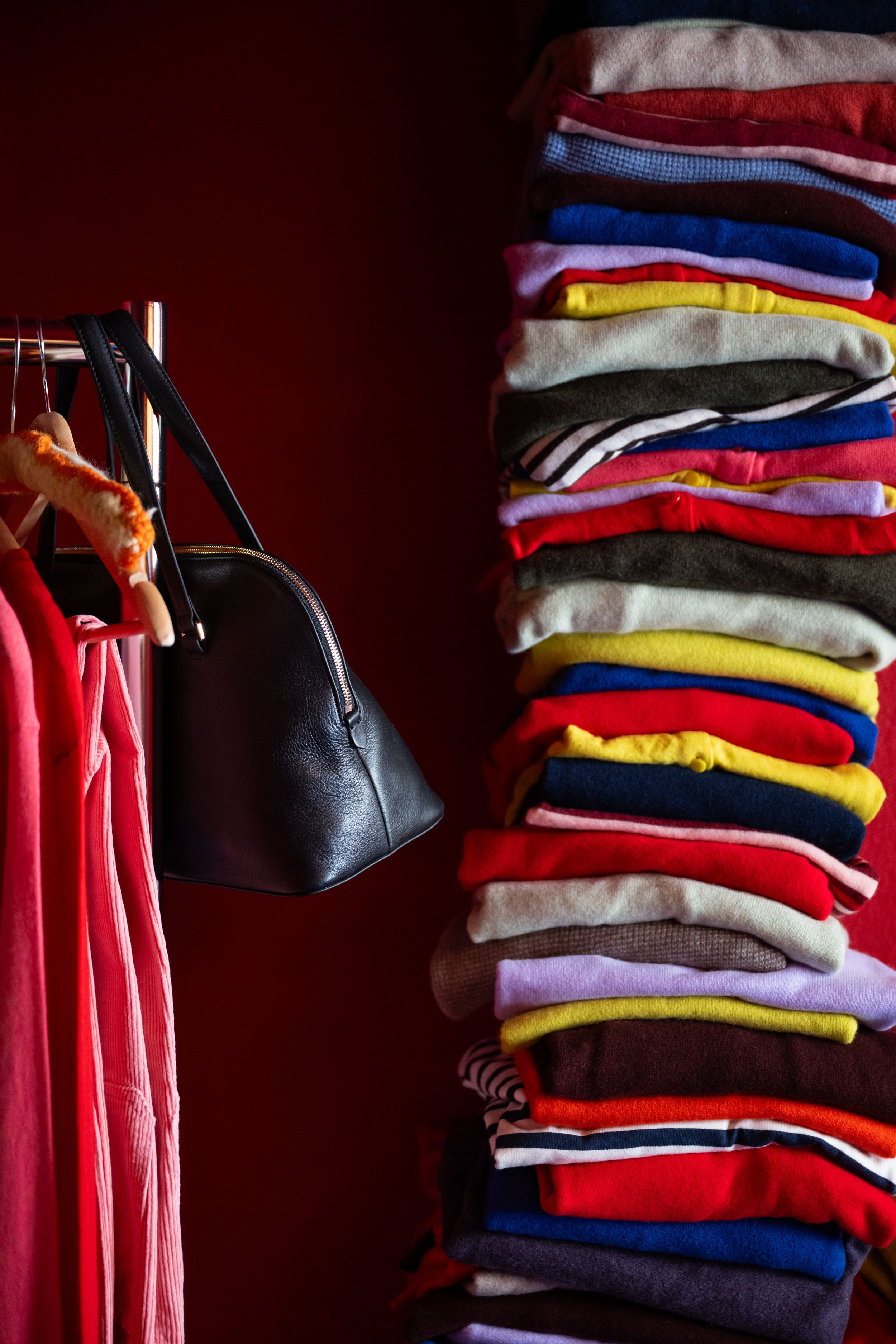 Colorful stack of folded sweaters next to hanging clothes and a black purse in a red closet.
