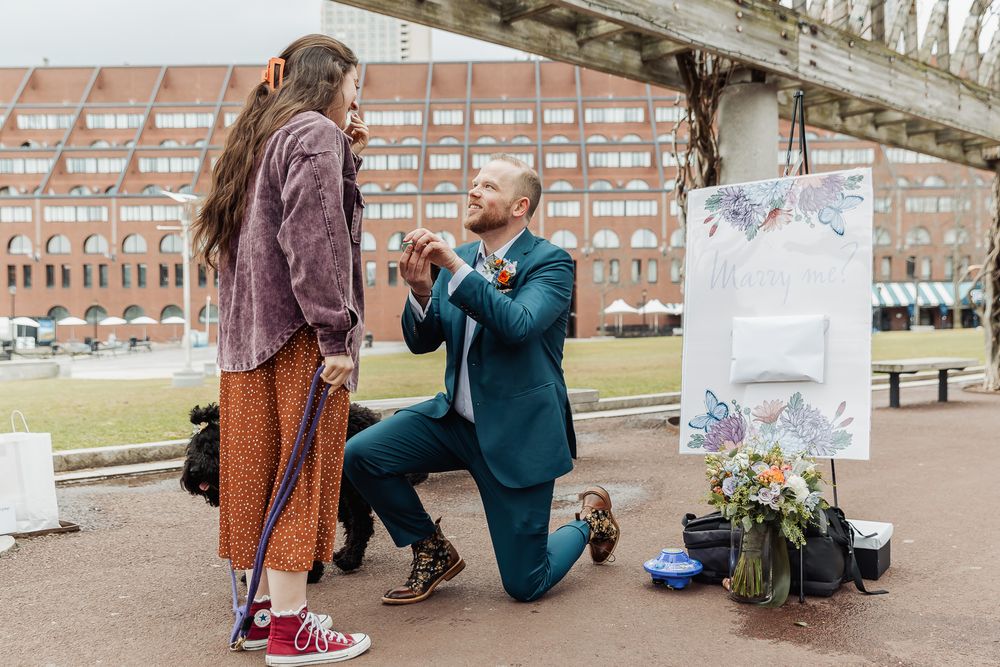 Man proposes on one knee to woman outdoors near floral arrangement and artwork, with historic building in background.