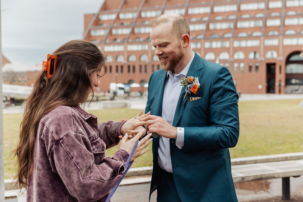 A smiling groom in a teal suit exchanges rings with a woman in a purple jacket outdoors.