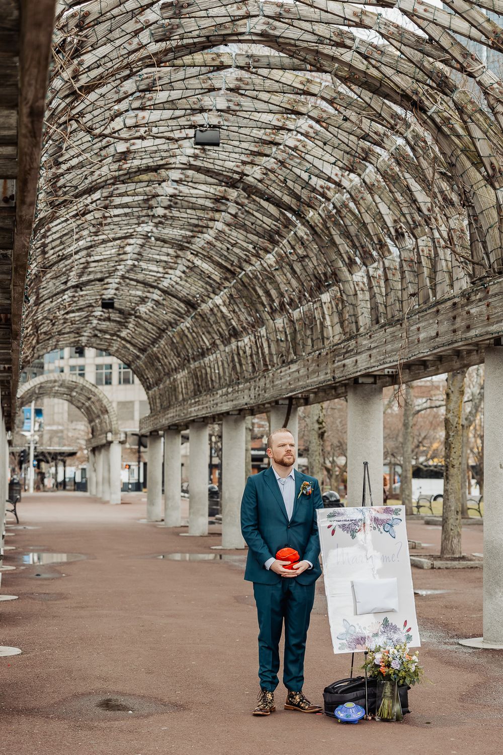 Groom in teal suit holds red boutonniere under stone arch pergola at outdoor wedding venue.