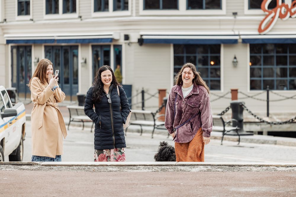 Three women smiling and laughing outdoors near a waterfront restaurant on a cloudy day.