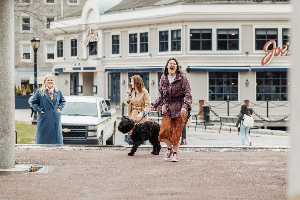 Three women walk a black curly dog in an urban plaza with a waterfront restaurant in the background.
