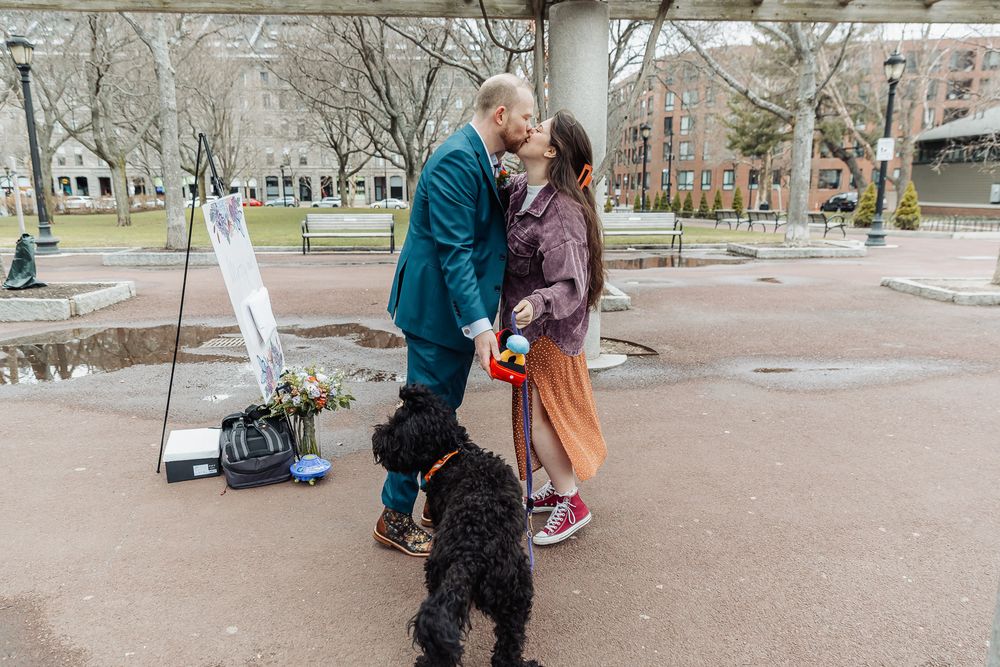 Couple sharing a kiss outdoors in a park with a black dog nearby, surrounded by bare trees and a puddle.