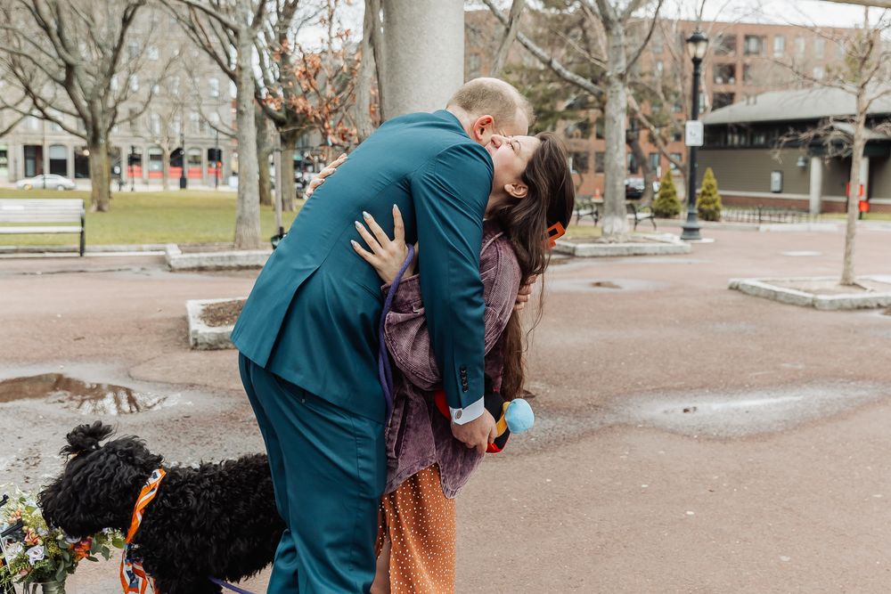 Couple sharing a romantic kiss outdoors in a city park, wearing colorful formal attire, with a black dog nearby.
