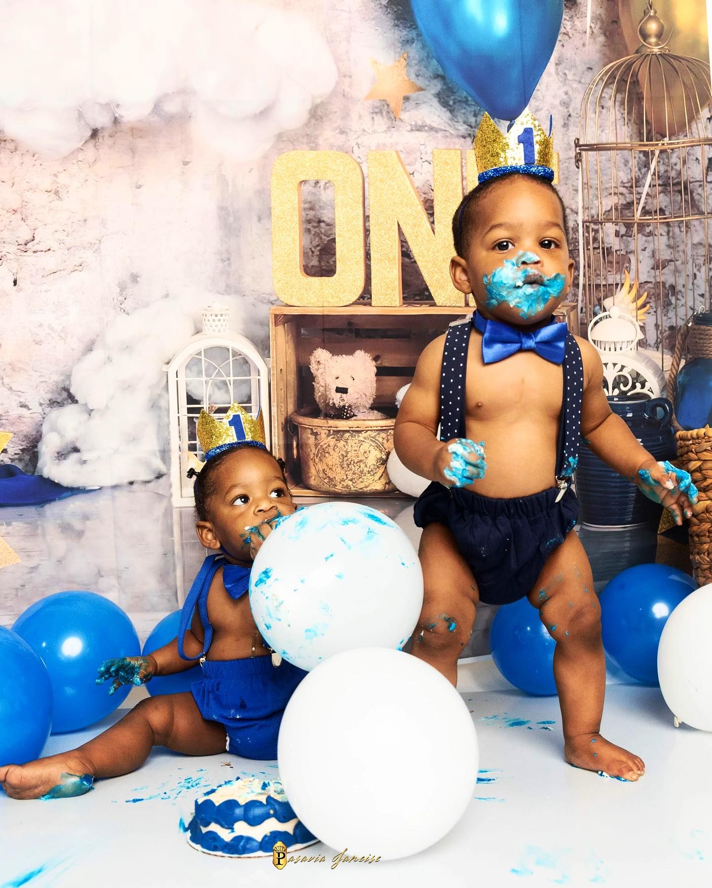 Two baby boys celebrate their first birthday with blue balloons, cake smash, and matching royal blue outfits with bow ties.