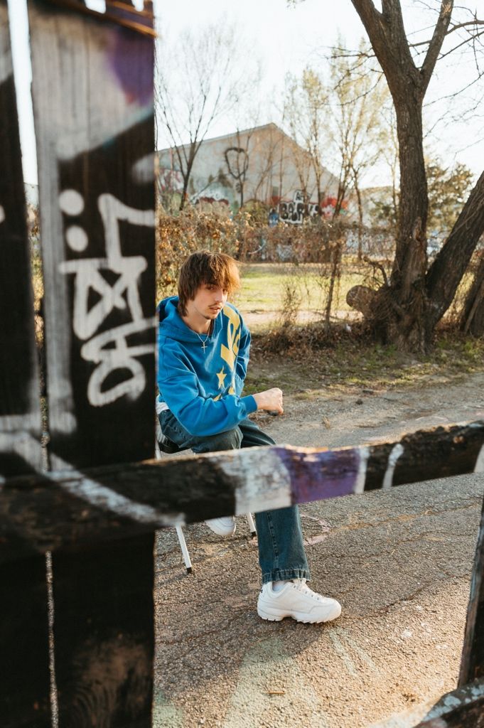 A teenage boy in a blue hoodie and jeans sits on a ledge near graffiti-covered walls in an urban outdoor setting.