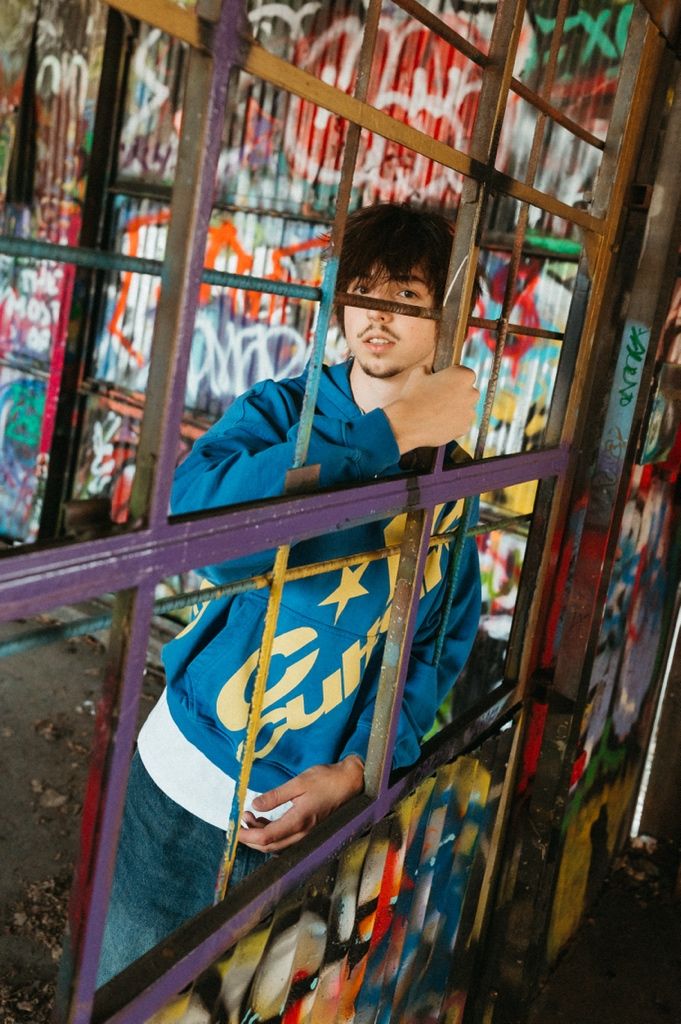 Young person in blue graphic hoodie poses behind a colorful graffiti-covered metal gate structure outdoors.
