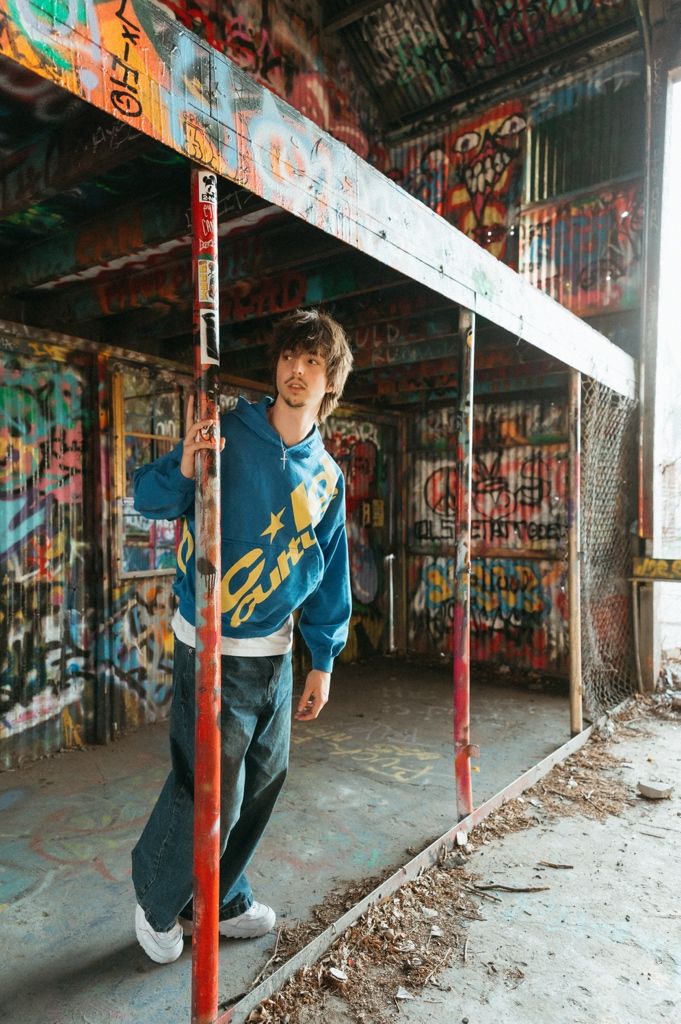Young man in blue hoodie and jeans poses in graffiti-covered abandoned building with colorful street art walls.