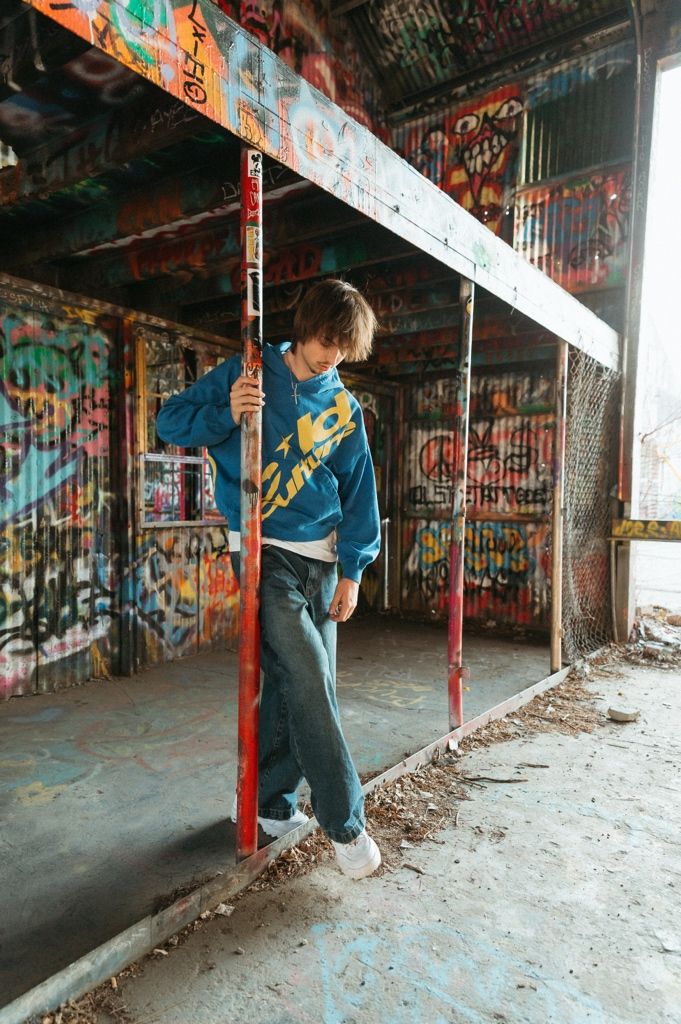 Young man in blue graphic hoodie holds red pole in vibrant graffiti-filled urban abandoned space.