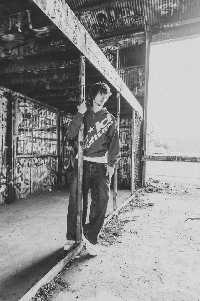 Black and white photo of a young man leaning on a metal pole in an abandoned graffiti-covered building.