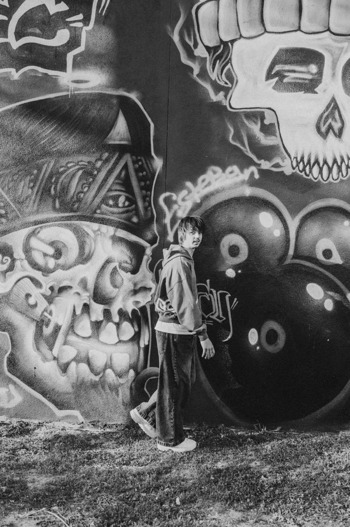 Black and white photo of a person standing in front of a large graffiti mural featuring skulls and bold street art.